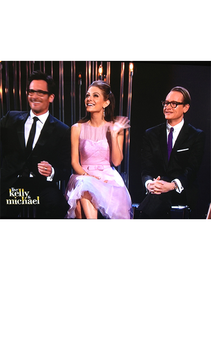 TV screen shot of 2 men in black suits and a woman in a lilac dress sitting